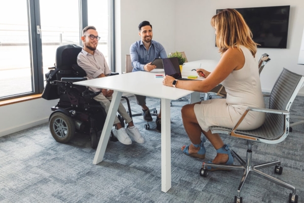 A woman with shoulder length blond hair wearing a white dress sits with her back to us at a desk in an office. Sat across the desk are two white men; one is a wheelchair user wearing a light shirt and glasses, the other is non-disabled with dark hair